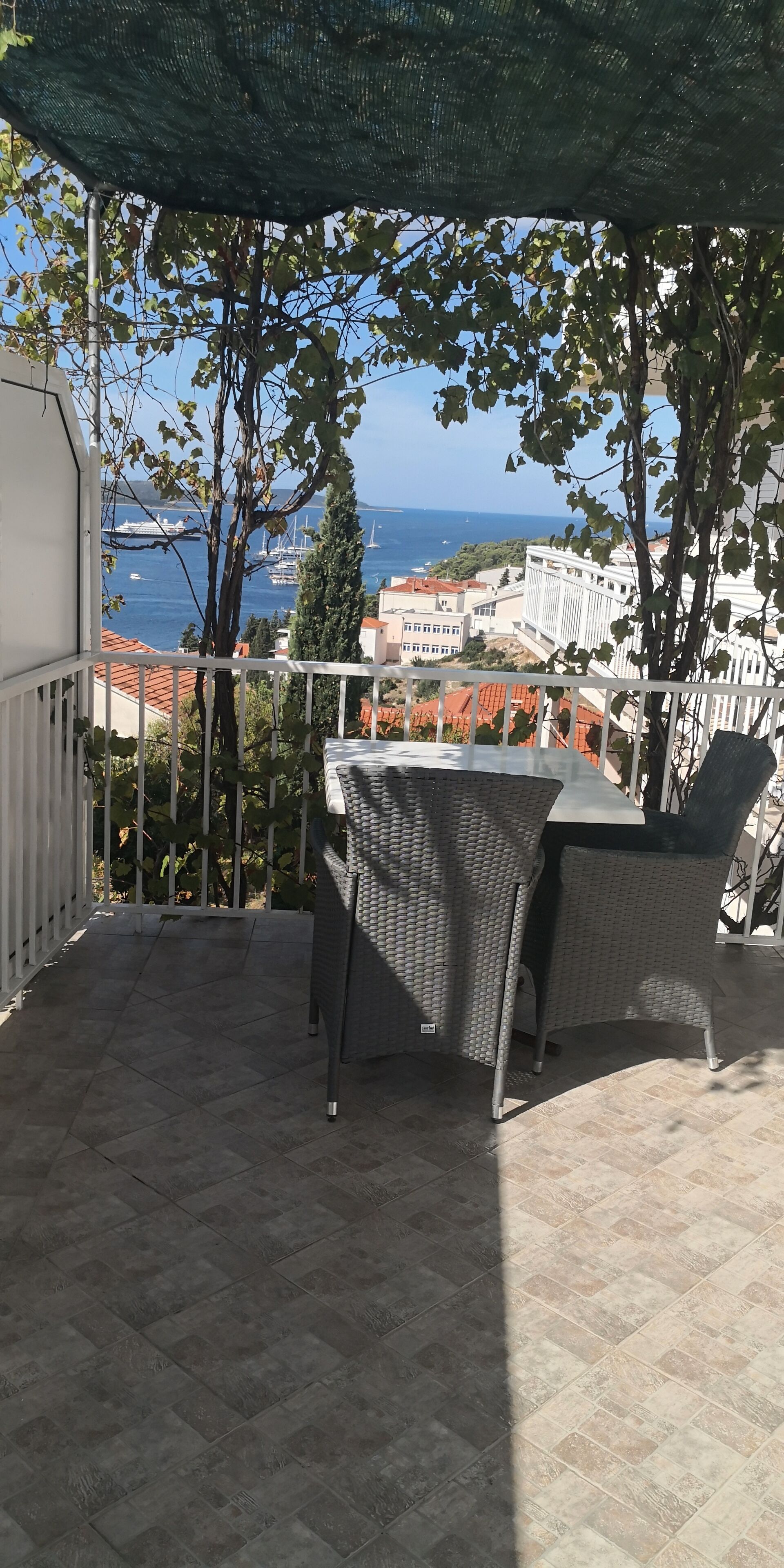 A balcony with a table and chairs, overlooking a coastal view with buildings and a clear sky.