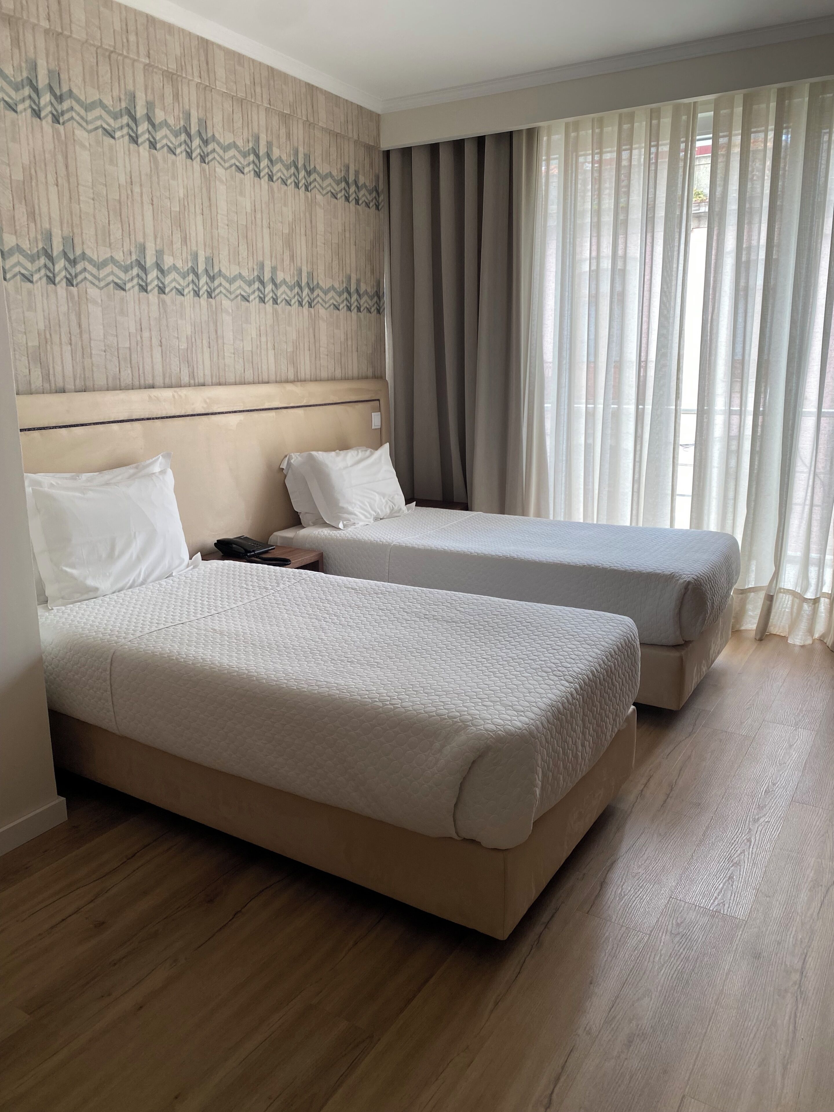 A hotel room with two beds, a wooden headboard, a patterned wall, and a view through sheer curtains.