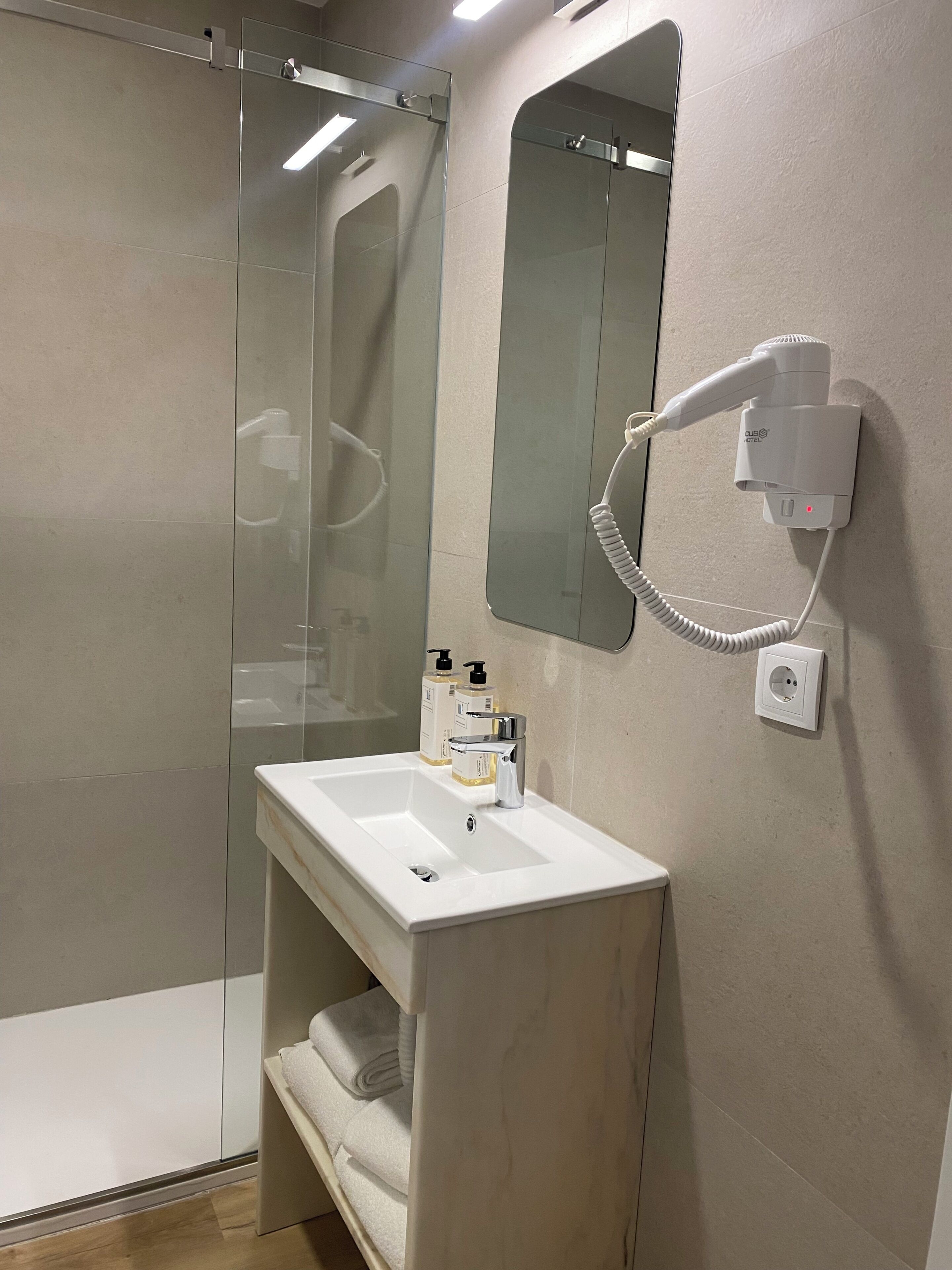 Une salle de bain avec une douche à parois de verre, un lavabo avec un miroir au-dessus et un sèche-cheveux branché.