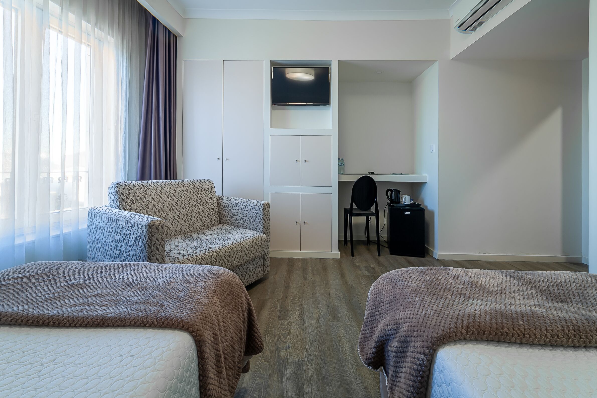 Quarto de hotel com duas camas, um sofá, uma escrivaninha com cadeira, televisão e guarda-roupa.