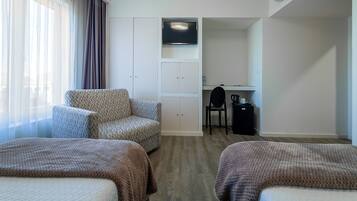 Quarto de hotel com duas camas, um sofá, uma escrivaninha com cadeira, televisão e guarda-roupa.