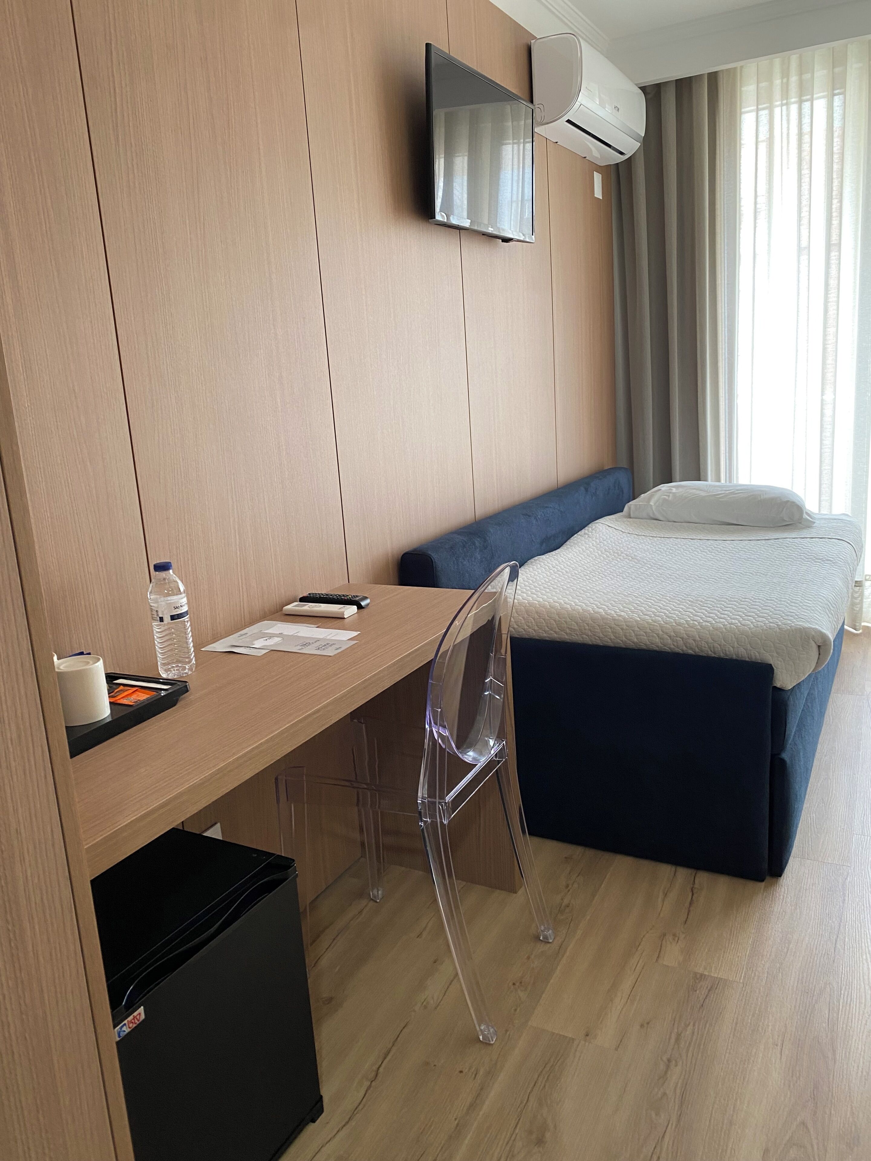 Une chambre d’hôtel avec un bureau en bois, une chaise transparente, un lit avec une tête de lit bleue, un téléviseur fixé au mur et un appareil de climatisation.