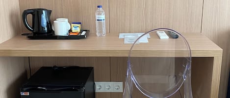 A wooden desk with a black coffee maker, a water bottle, a mug, and a remote control. A clear plastic chair is in front of the desk.