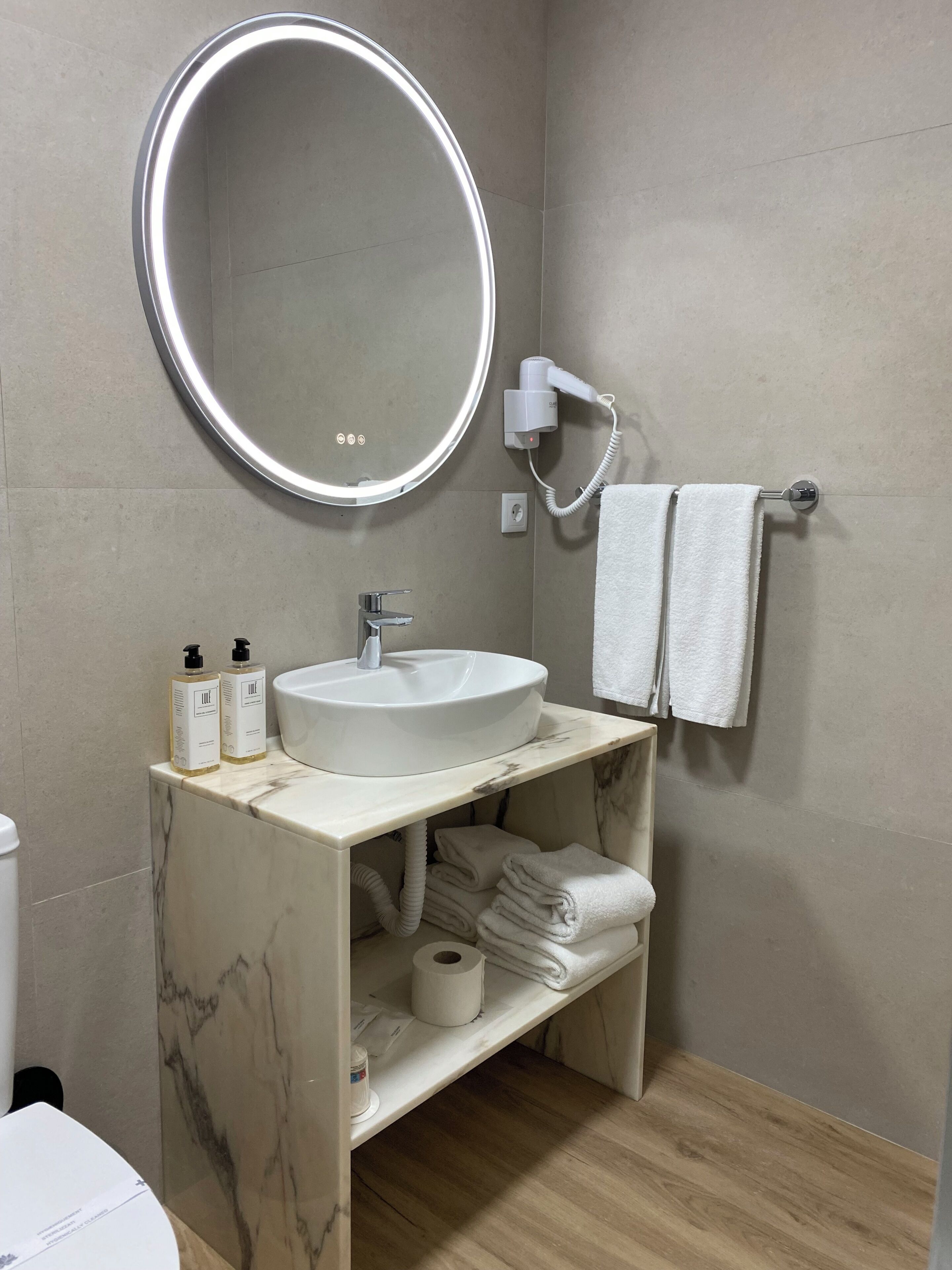 A bathroom with a round mirror, white sink, two bottles of lotion, a towel rack with two towels, and a toilet.