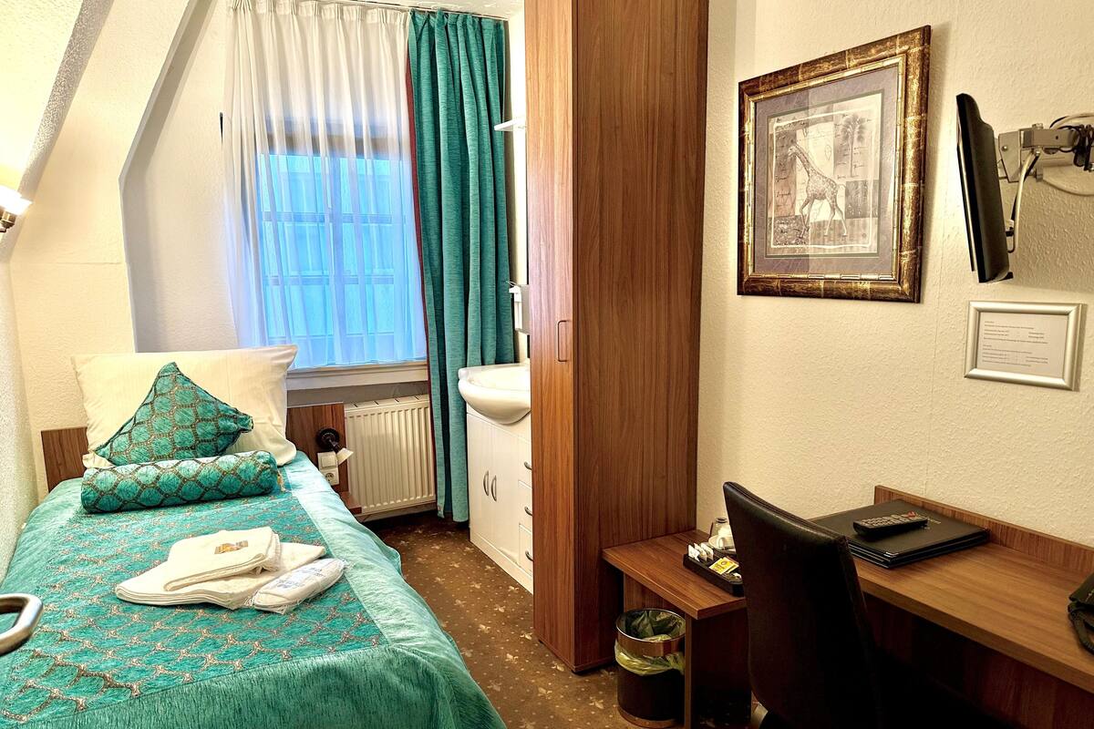 Habitación de hotel con cama, escritorio, radiador, ventana con cortinas, un cuadro enmarcado y un cubo de basura.