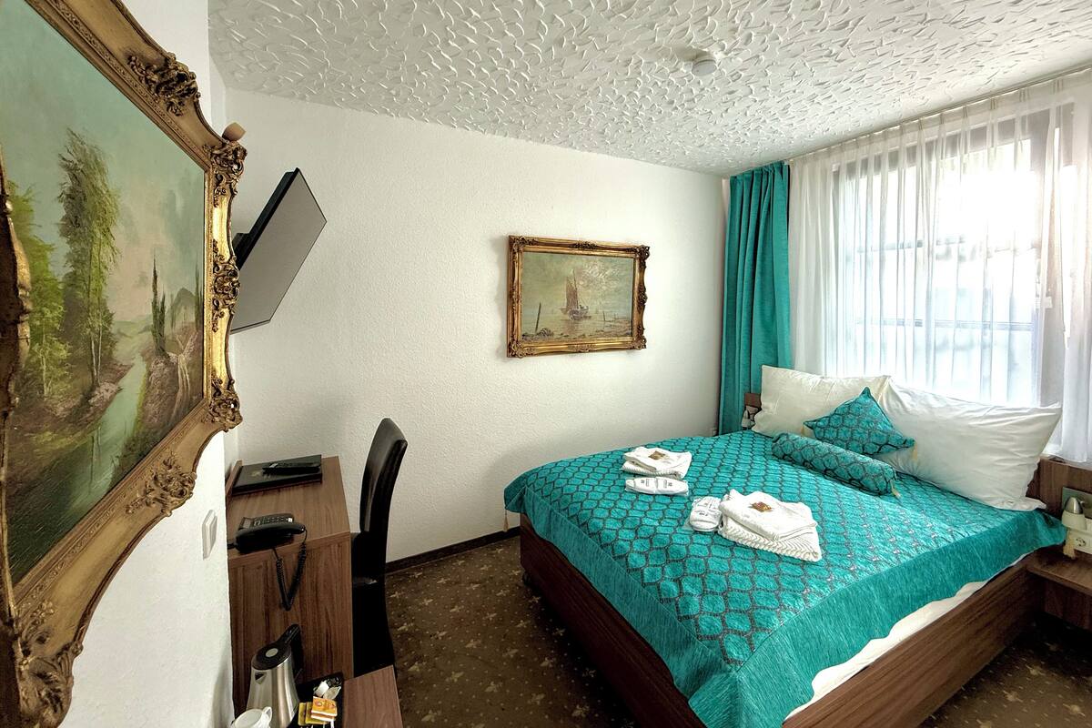 Habitación de hotel con cama, escritorio con teléfono, una silla, un cuadro en la pared y una ventana con cortinas.