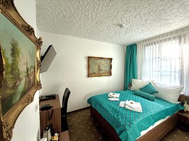 Quarto de hotel com cama, escrivaninha com telefone, cadeira, quadro na parede e janela com cortinas.