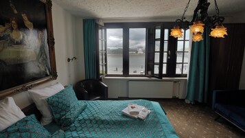 A room with a bed, a chair, a radiator, a window with a view of a body of water, and a painting on the wall.