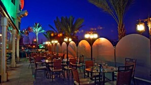 An outdoor dining area with tables and chairs, palm trees, and a view of the city skyline at night.