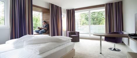 Habitación de hotel con una cama grande, una mesita pequeña, una silla y vistas a árboles por las ventanas.