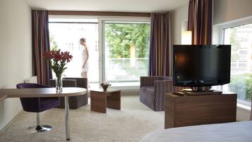 Quarto de hotel com cama, televisão, mesa de jantar, cadeira, pequena mesa com planta e vista para uma varanda.