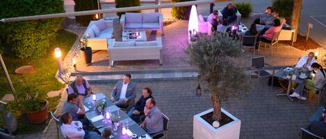 An outdoor patio with tables and chairs, surrounded by potted plants and a large tree.