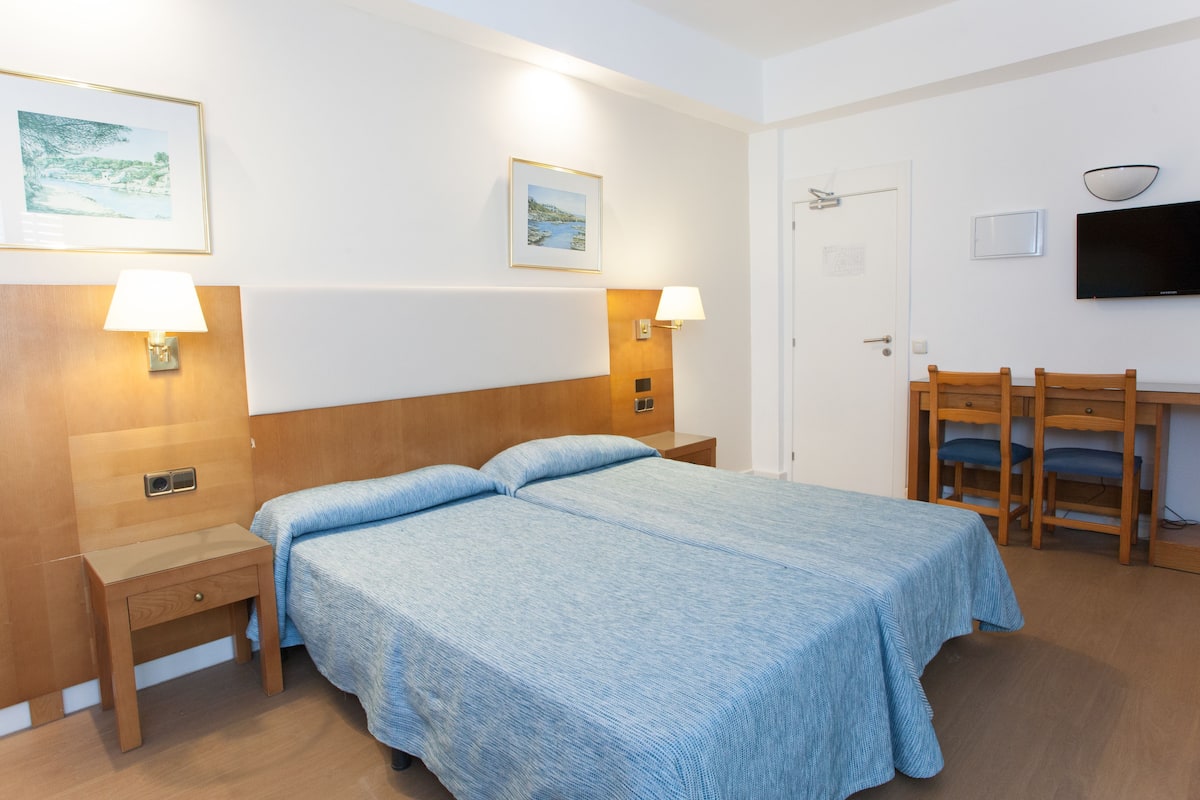 A hotel room with a bed, two bedside tables with lamps, a desk with a chair, and a TV on the wall.