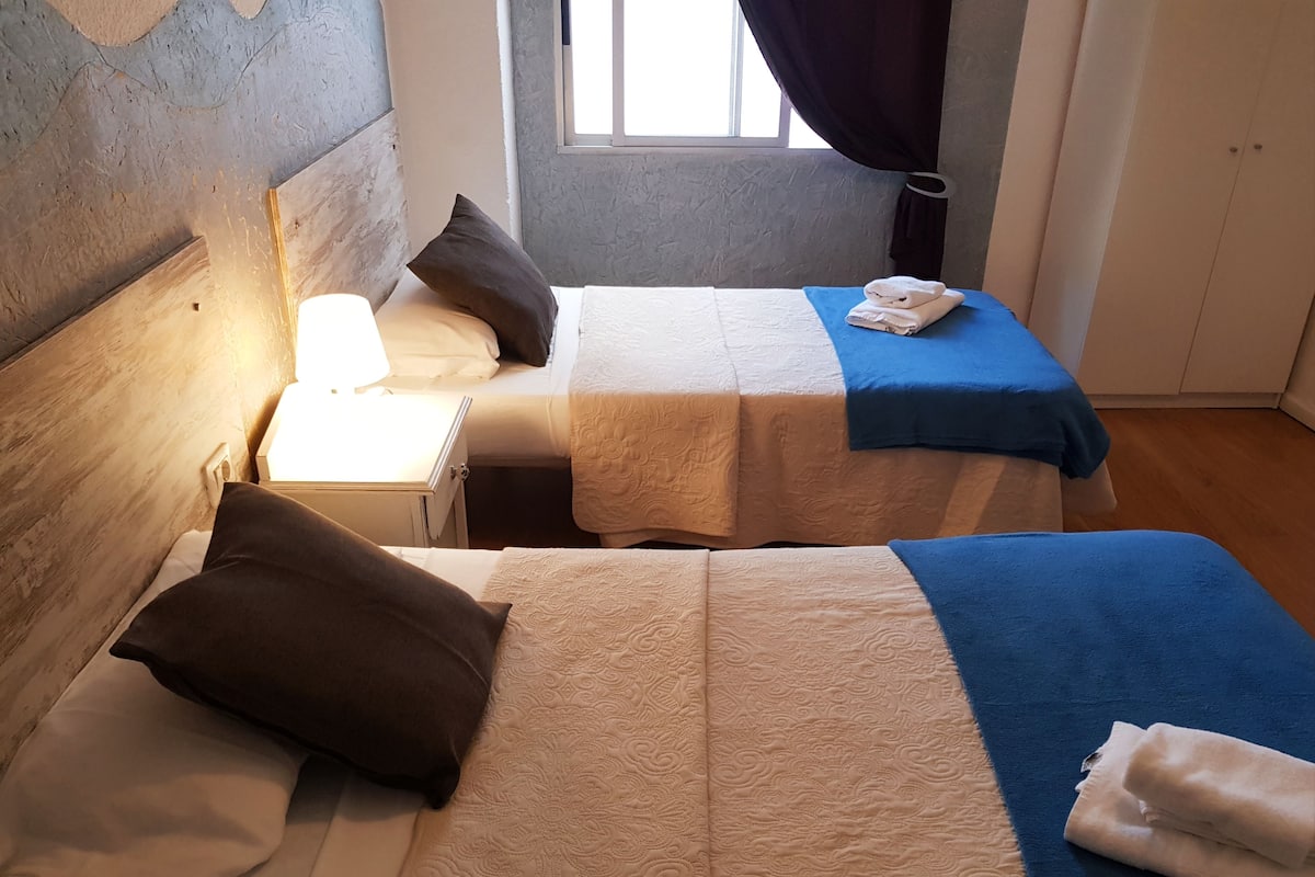 Une chambre avec deux lits, chacun doté d’une tête de lit, d’une table de chevet, d’une lampe et d’une fenêtre avec des rideaux.