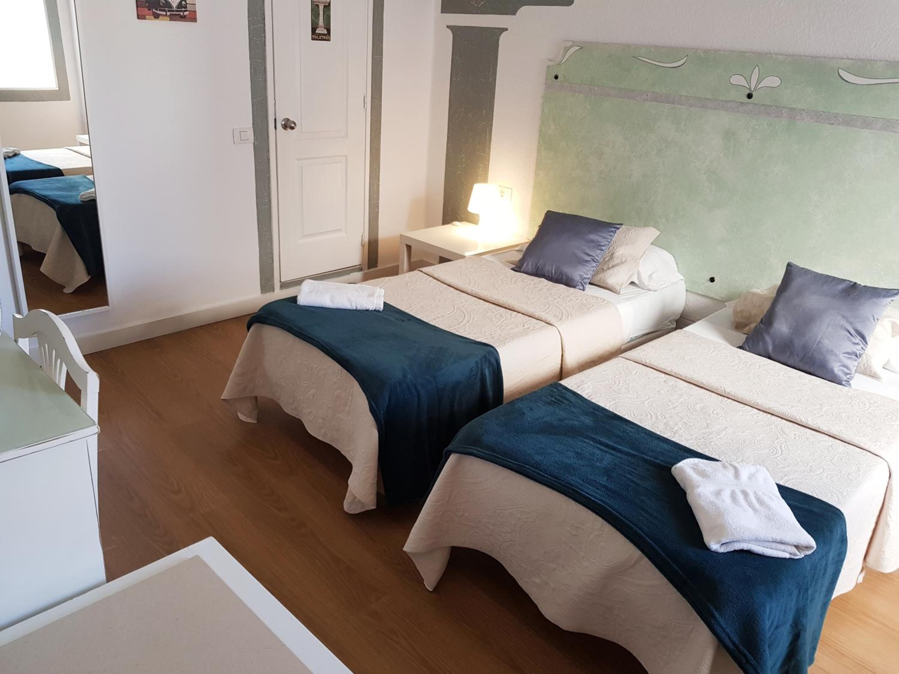 A hotel room with two beds, a small bedside table with a lamp, a mirror, and a wooden floor.