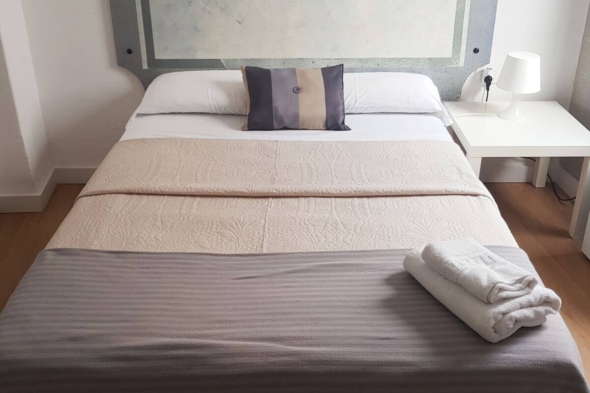 Un lit bien fait, avec une couvre-lit à motifs, un oreiller et une serviette pliée, posés sur un plancher en bois.