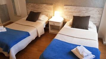 Ein Hotelzimmer mit zwei Betten, jedes mit einer blauen Decke und weißen Kissen. Dazwischen steht ein Nachttisch mit einer Lampe.
