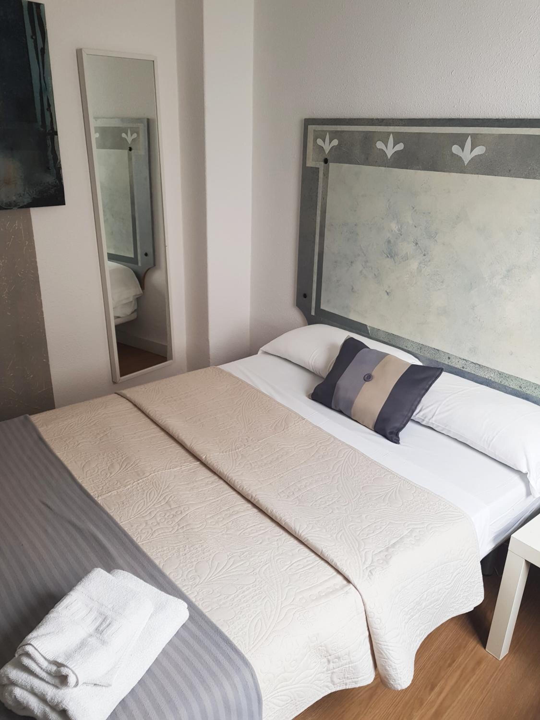 A double bed with a beige bedspread and two pillows, a bedside table, and a mirror on the wall.