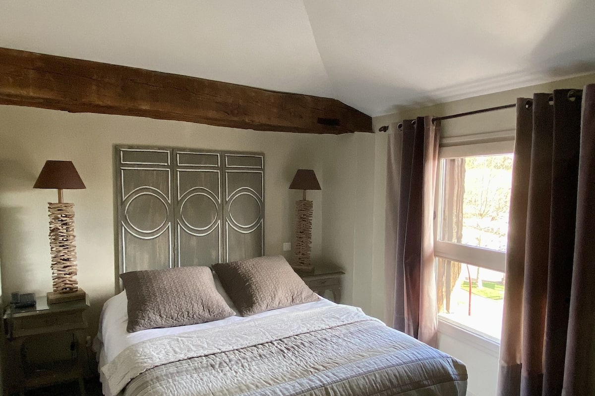 A bedroom with a large bed, two bedside tables with lamps, a wooden ceiling beam, and a window with curtains.