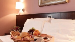 Una cama bien hecha con sábanas blancas, una mesita de noche con una lámpara, y una bandeja de desayuno con pan, fruta y una taza de café.