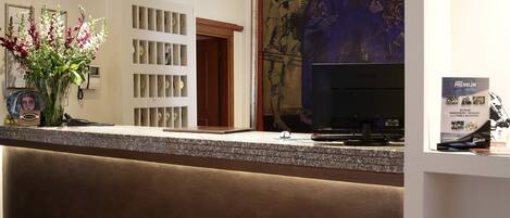 Zona de recepción de un hotel con mostrador, televisor, arreglo floral y un cuadro grande en la pared.