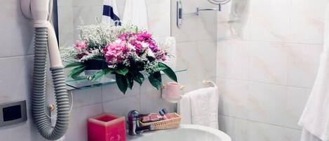 Un baño con lavamanos blanco, secador de pelo, espejo y estante con flores y artículos de tocador.