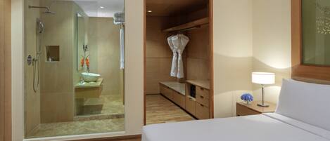 A hotel room with a walk-in closet, a separate bathroom with a glass shower enclosure, and a bedside table with a lamp.