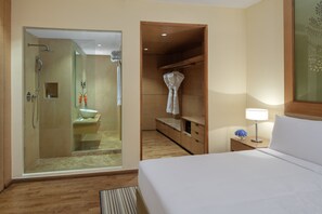 A hotel room with a walk-in closet, a separate bathroom with a glass shower enclosure, and a bedside table with a lamp. - Radisson Blu Hotel New Delhi Dwarka (New Delhi)