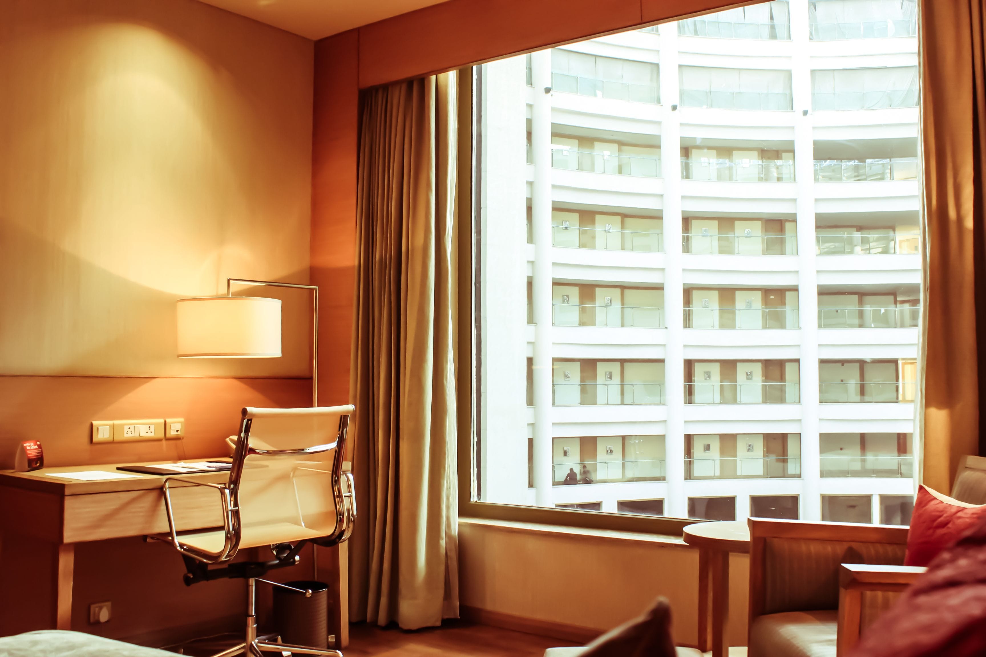 Quarto de hotel com escrivaninha, cadeira e uma janela grande que oferece vista para um arranha-céu.