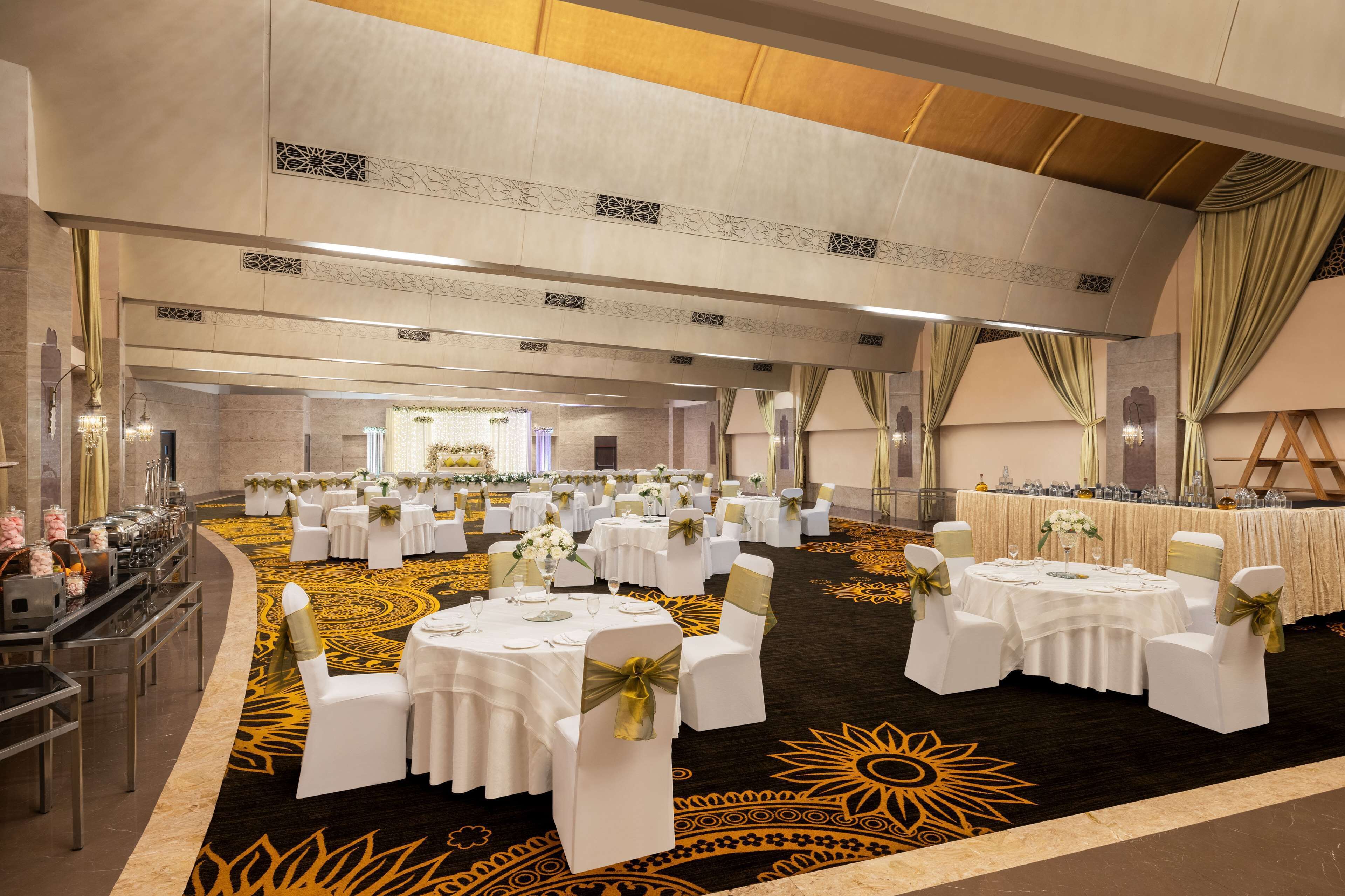 Une salle de banquet avec des tables rondes dressées pour le repas, des chaises blanches ornées de rubans dorés et une moquette à motifs solaires.
