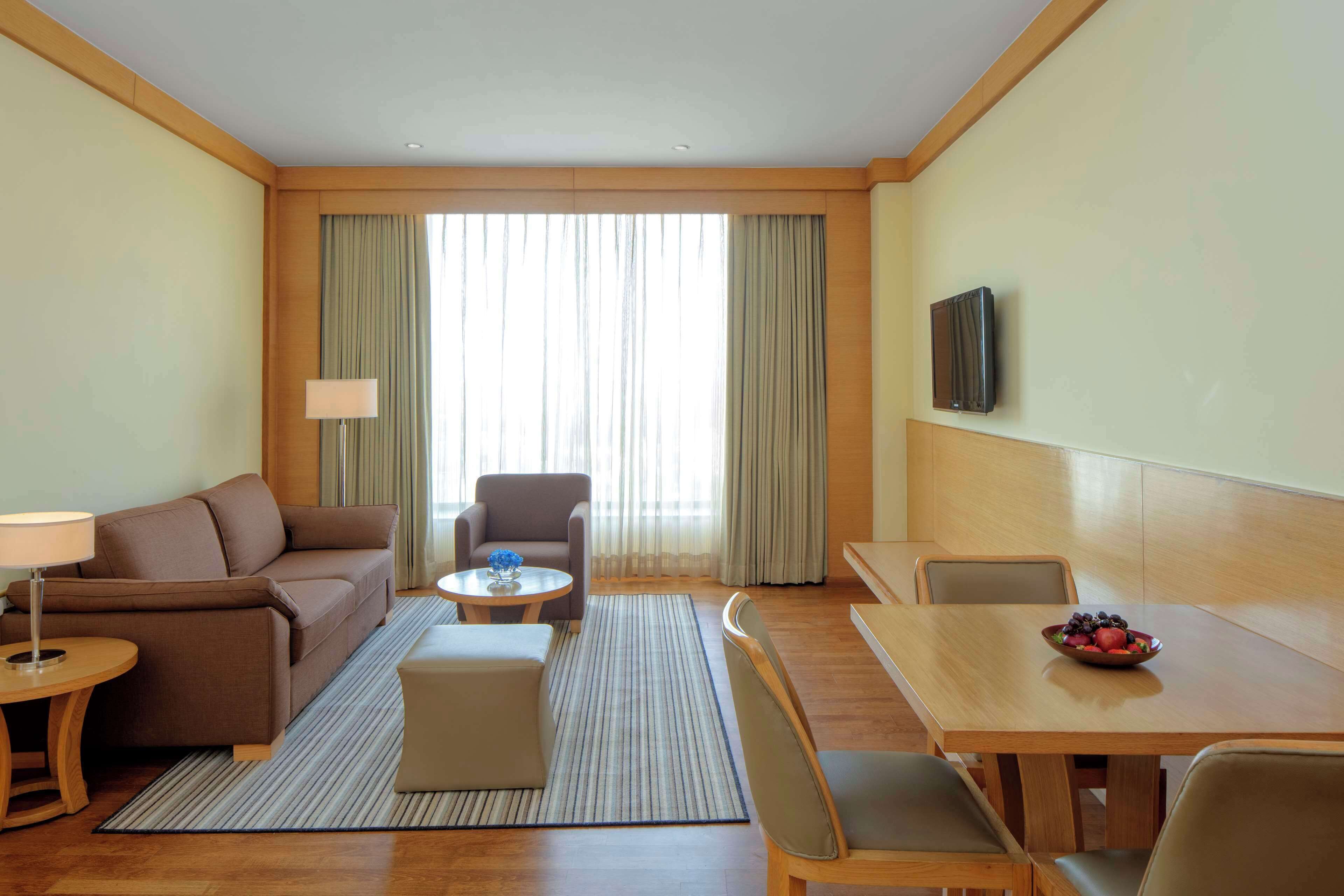 A hotel room with a sofa, armchair, small table, ottoman, dining table with chairs, and a TV mounted on the wall.