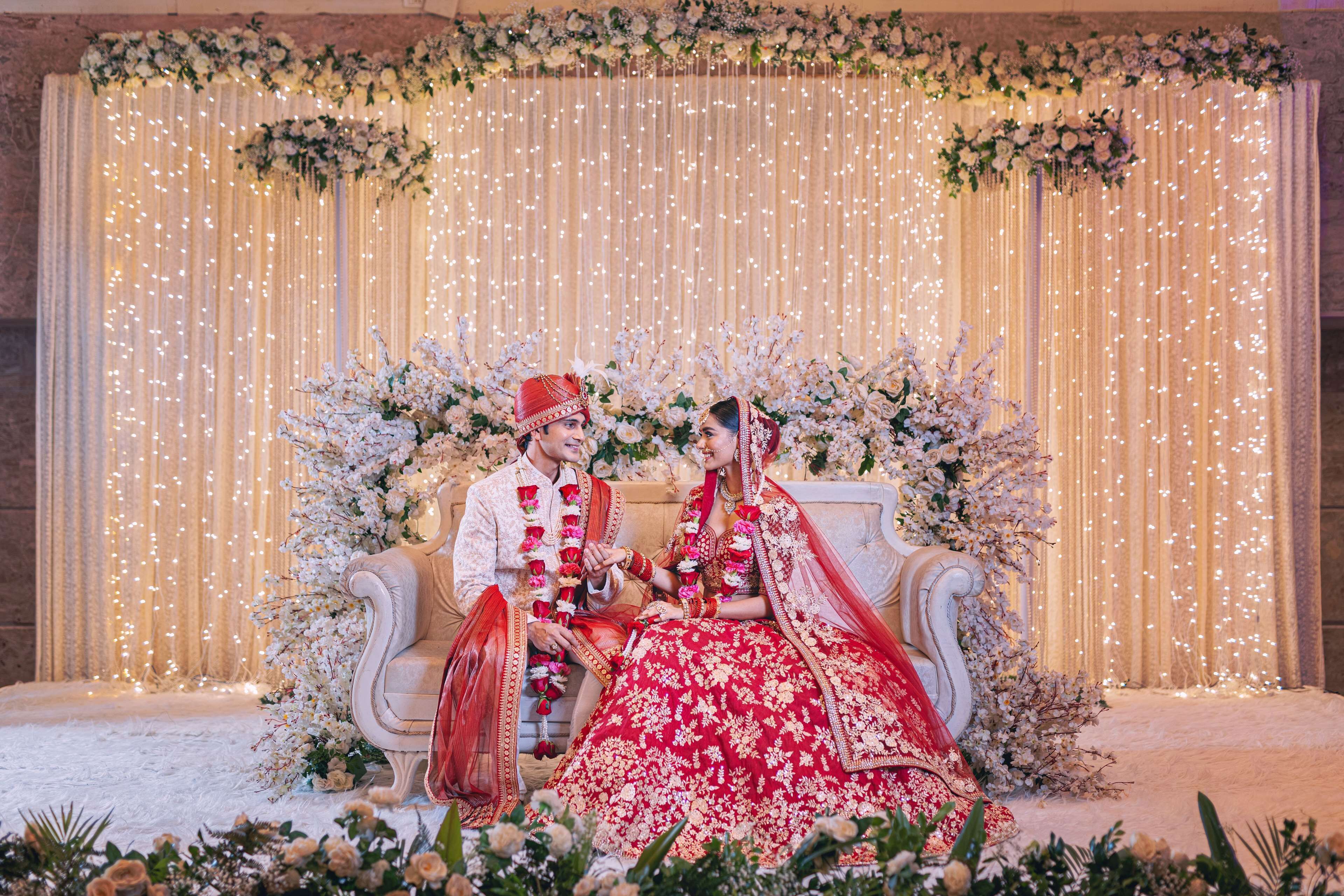 Una pareja sentada en un sofá ornamentado, con arreglos florales e iluminación de luces colgantes al fondo.