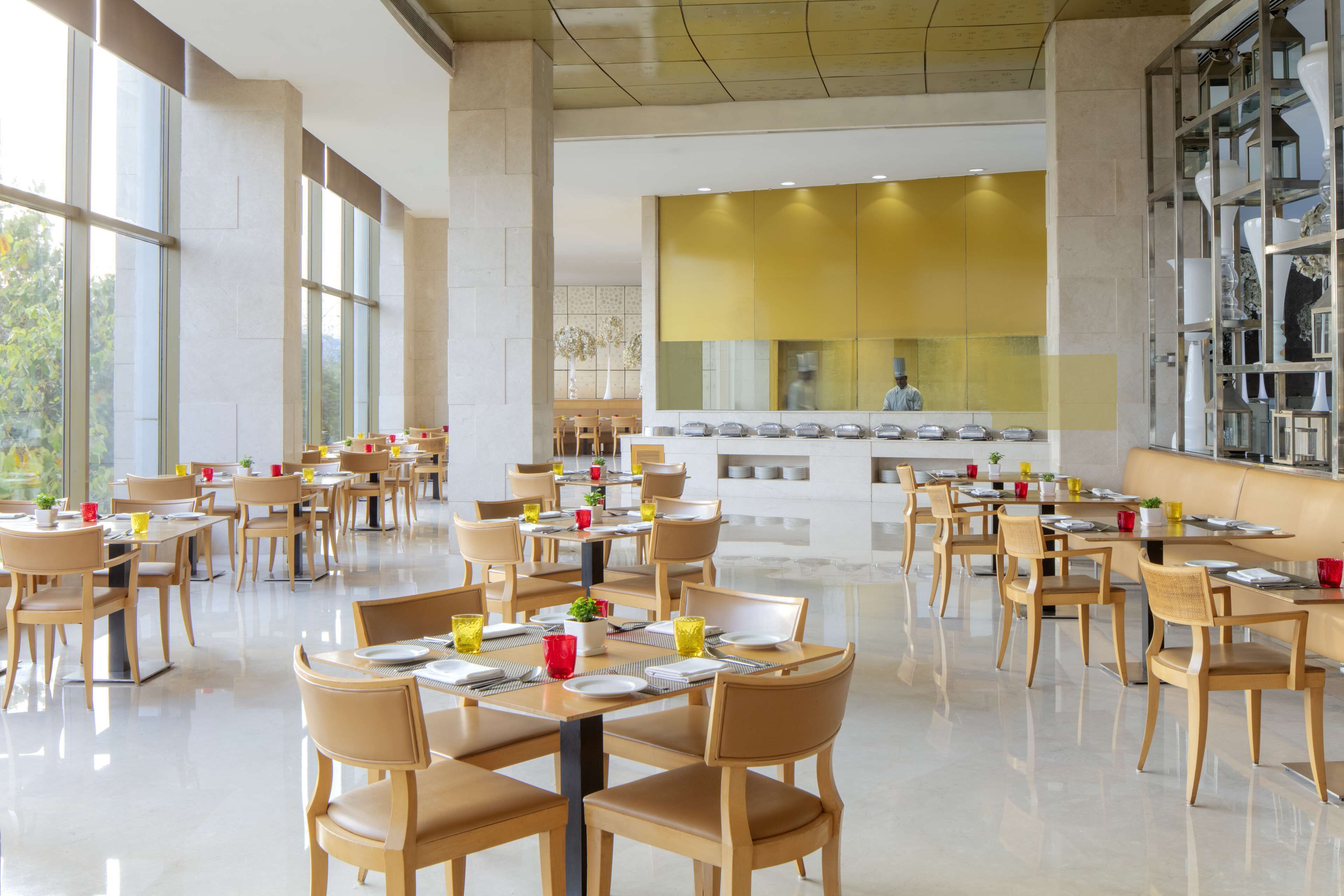 Un vaste espace de salle à manger avec plusieurs tables et chaises, de grandes fenêtres et, au fond, un coin cuisine moderne.