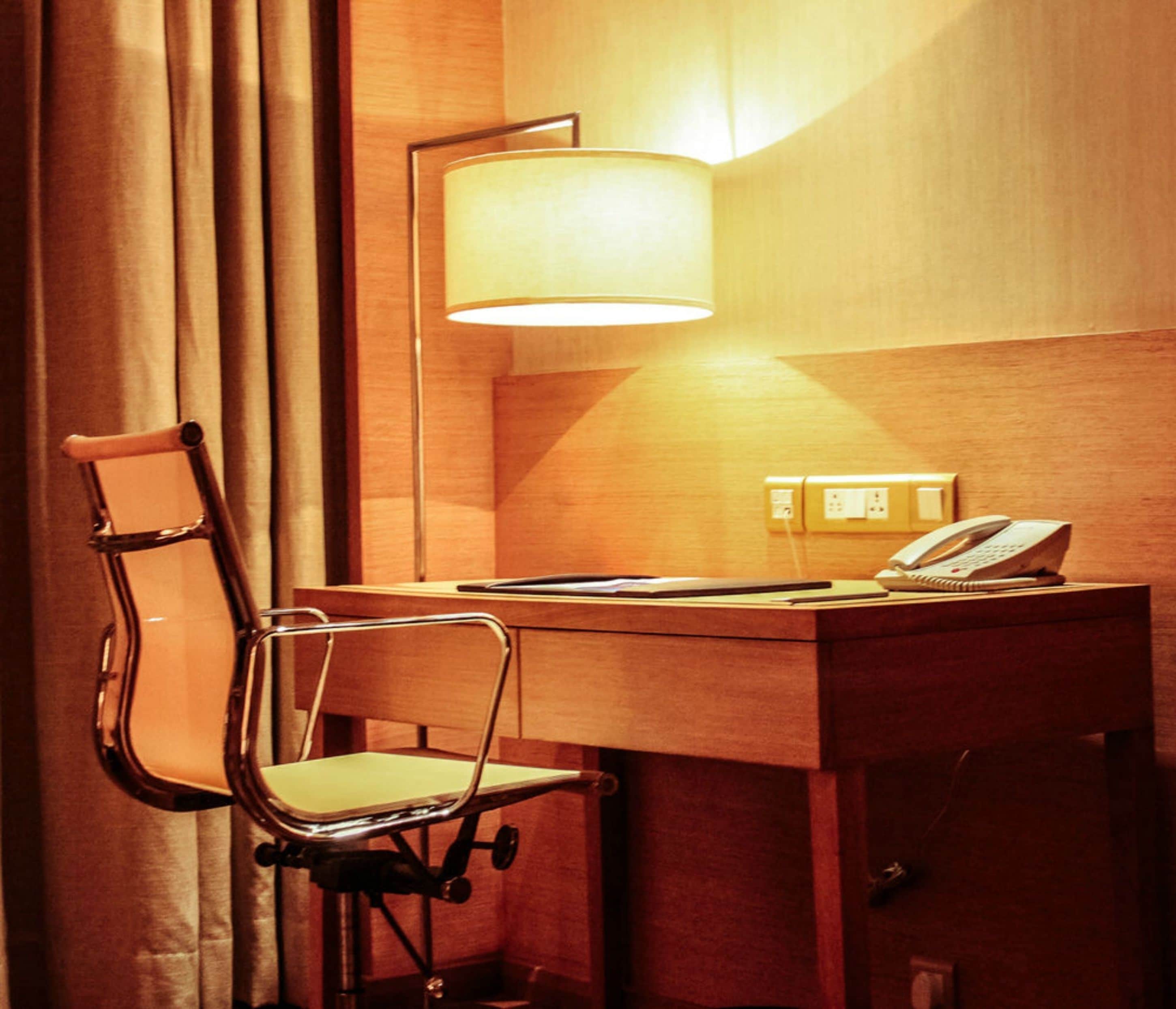 A hotel room with a wooden desk, a chair, a lamp, a telephone, and a power outlet.