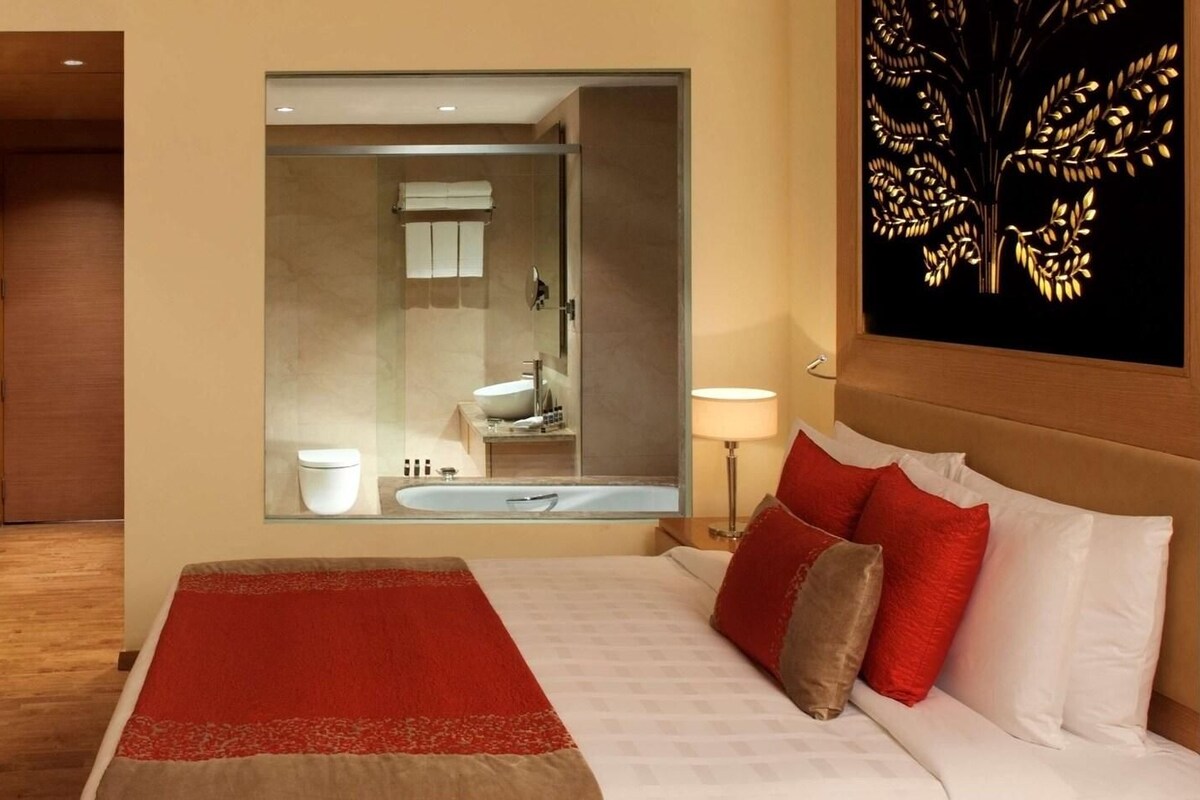 A hotel room with a bed, a nightstand, a dresser, and a bathroom visible through a glass partition.