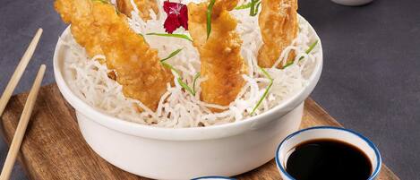 A bowl of fried items on rice with dipping sauces and a wooden board.