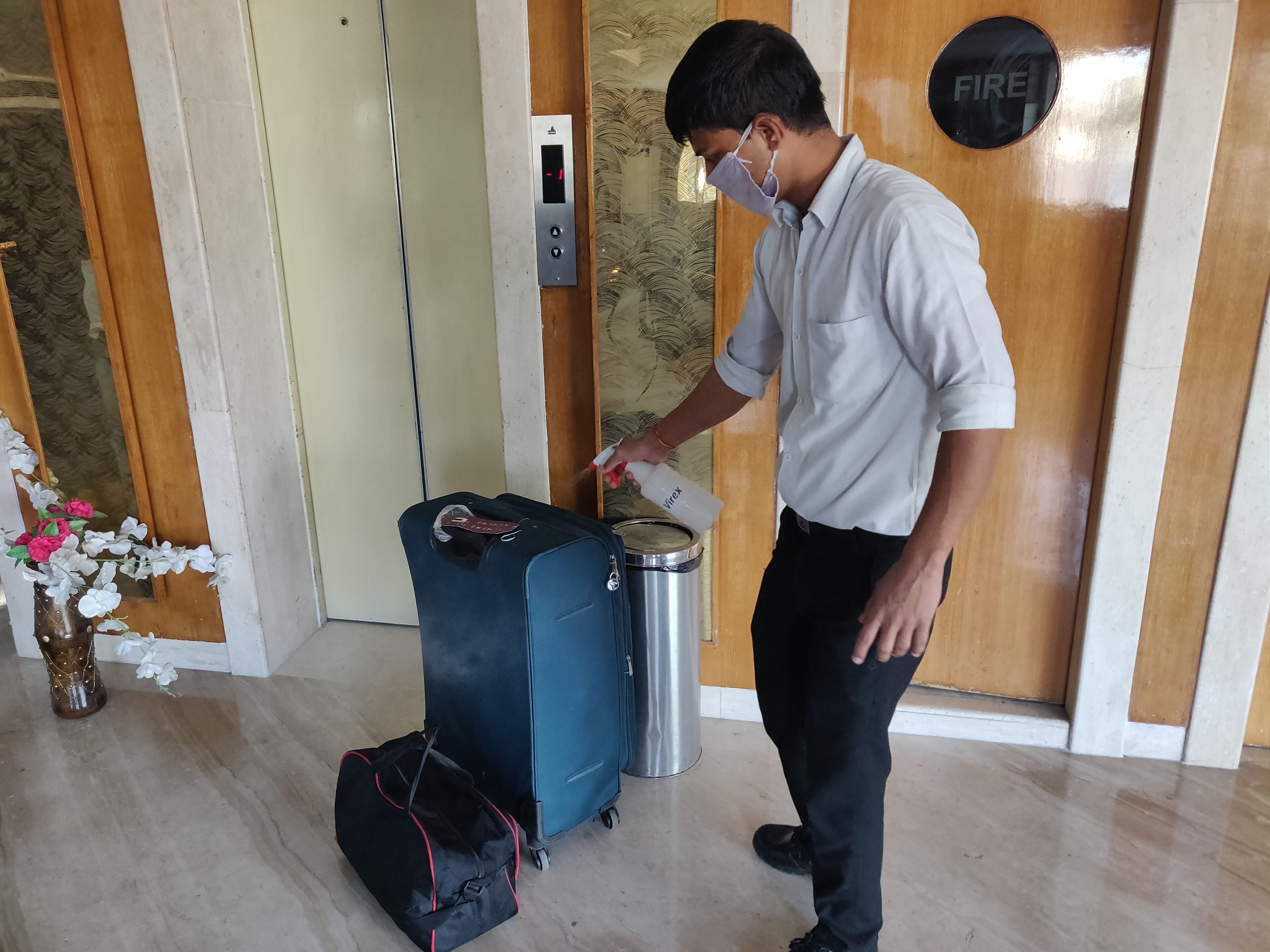 Une personne vêtue d’une chemise blanche et d’un pantalon noir manipule des bagages à proximité d’un détecteur d’incendie et d’un panneau de commande numérique pour ascenseur.