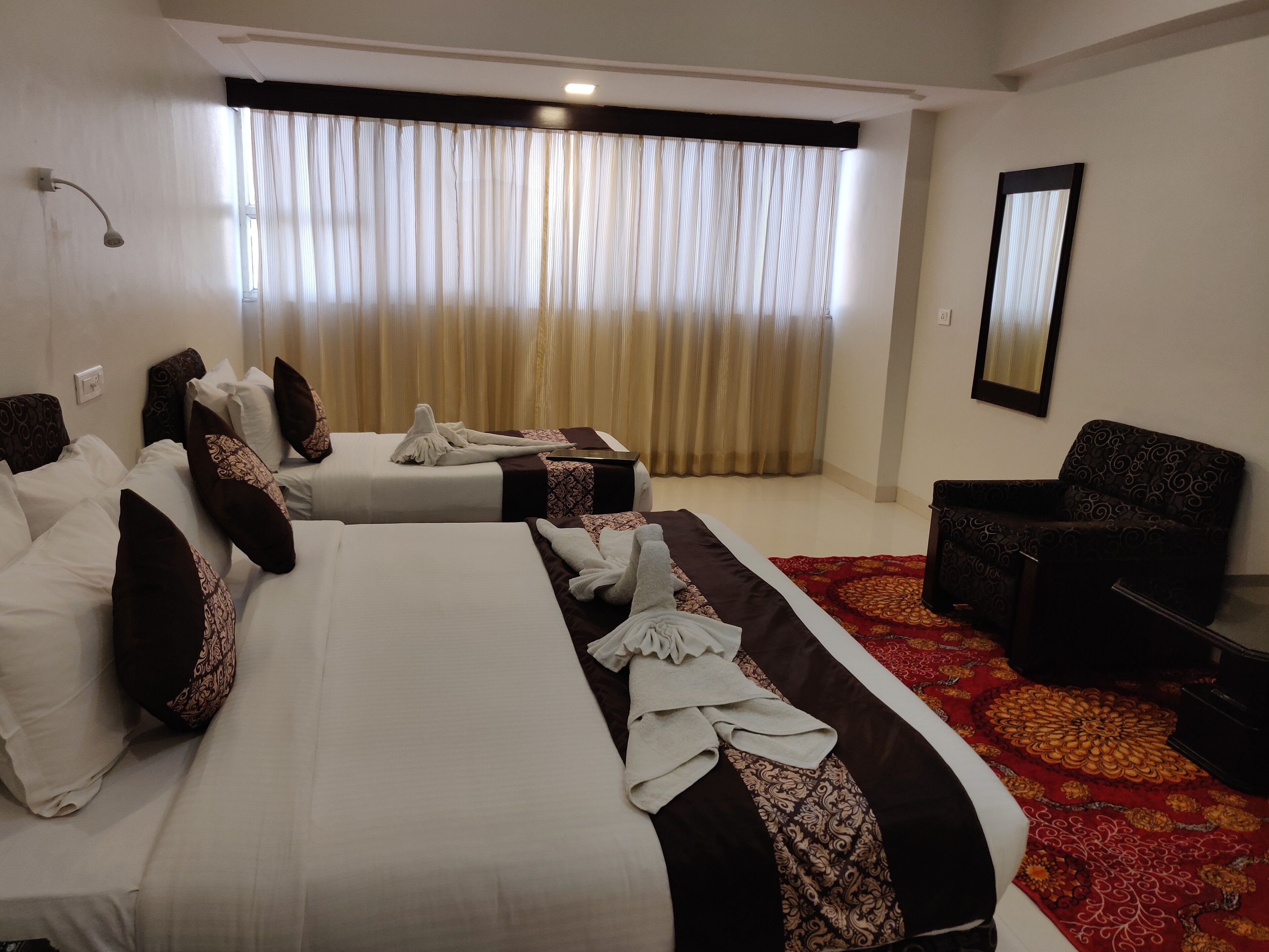 A hotel room with two beds, a sofa, a chair, a large window with curtains, and a mirror on the wall.