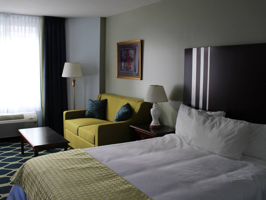 A hotel room with a bed, a yellow armchair, a small table, and a lamp.