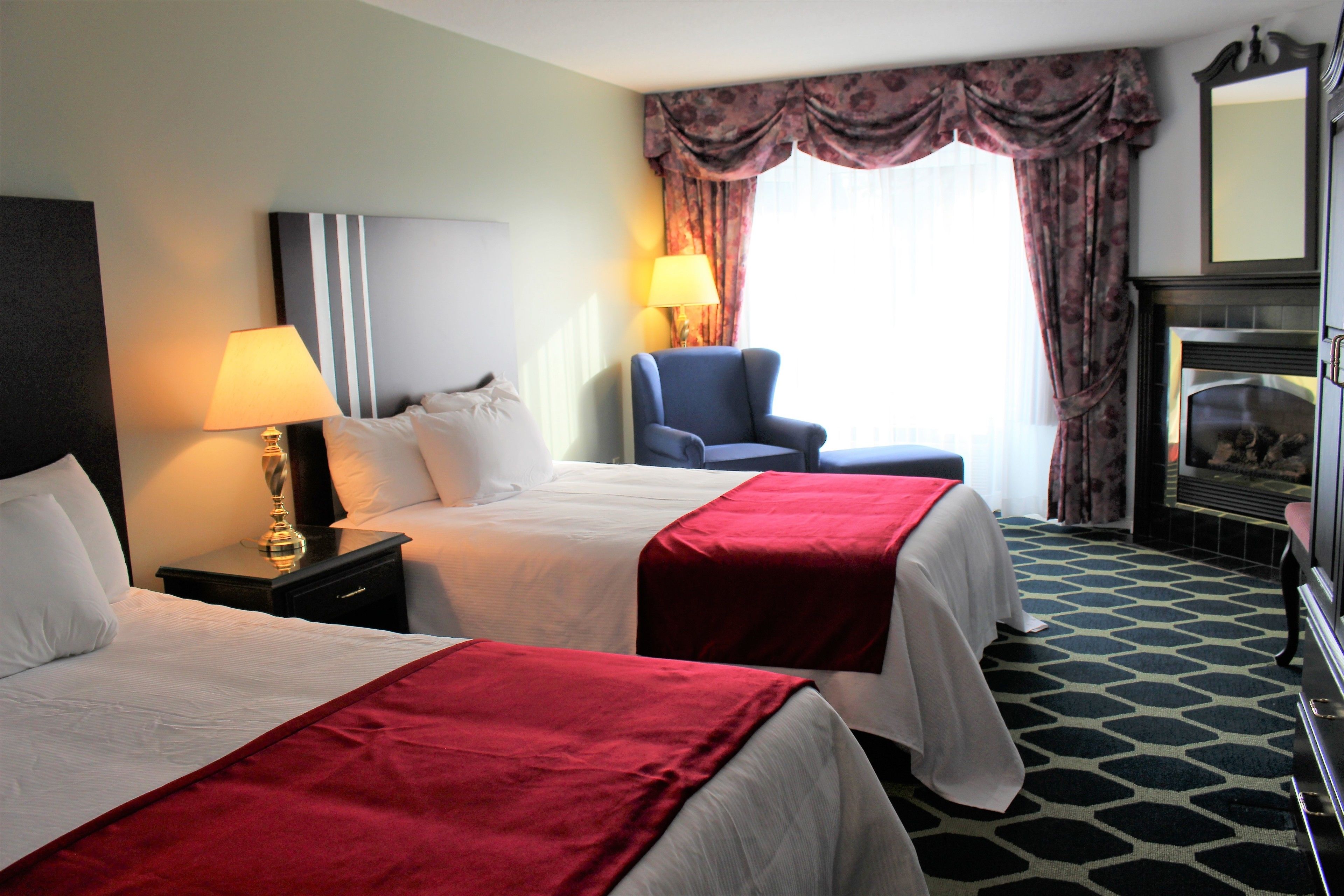 A hotel room with two beds, a nightstand with a lamp, a chair, a fireplace, and a large mirror.