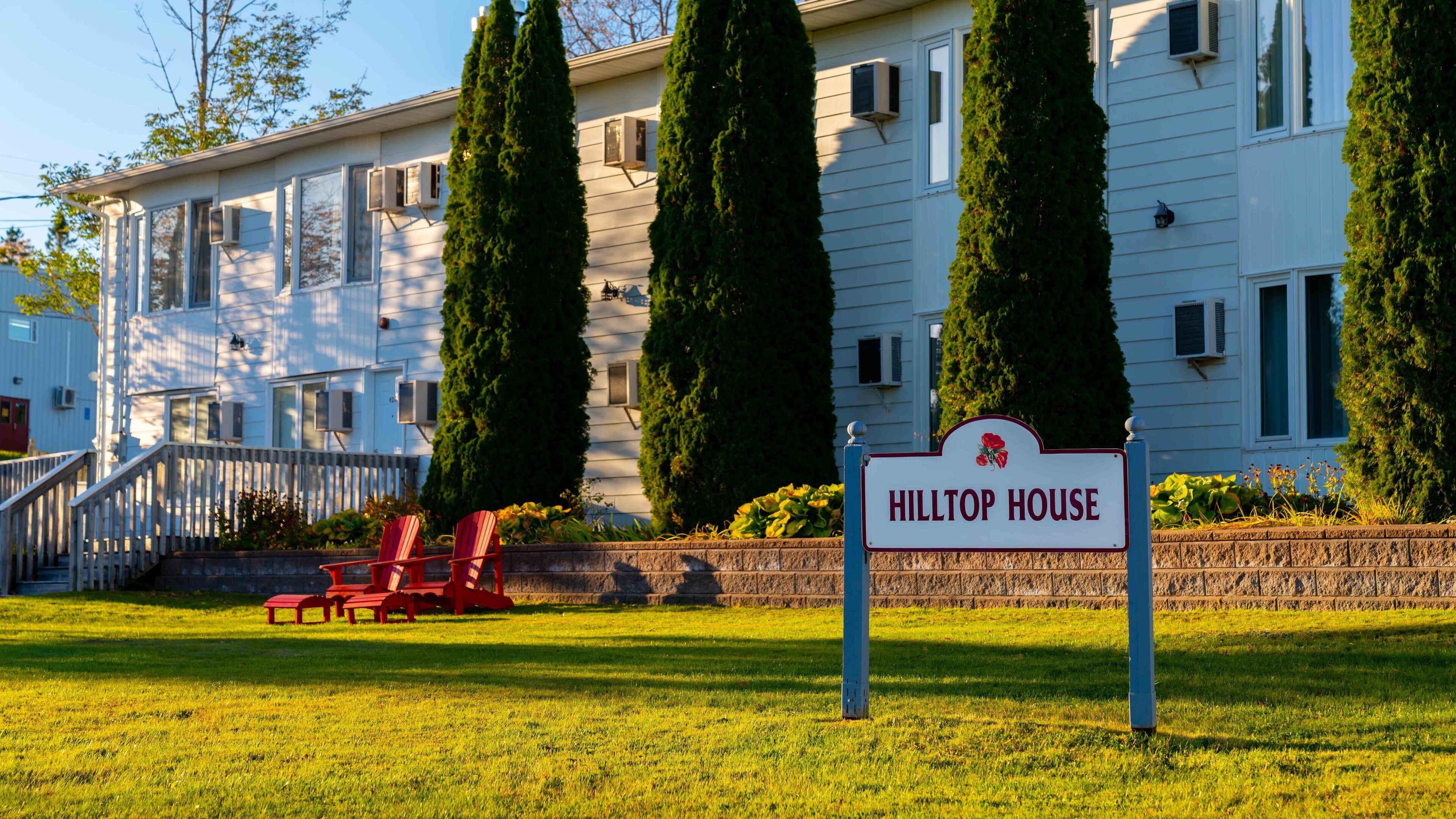 A sign for 'HILTOP HOUSE' with a staircase leading to a white building, surrounded by greenery and red chairs.