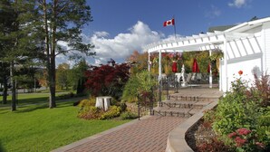 A white building with a Canadian flag, surrounded by trees and a garden with flowers and shrubs. - Auberge Gisele's Inn (Baddeck)