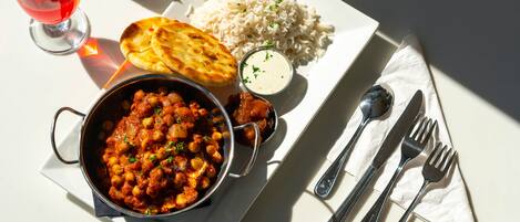 Refeição com arroz, pão naan e ensopado de grão-de-bico, servida num prato branco com talheres e um copo de bebida vermelha.