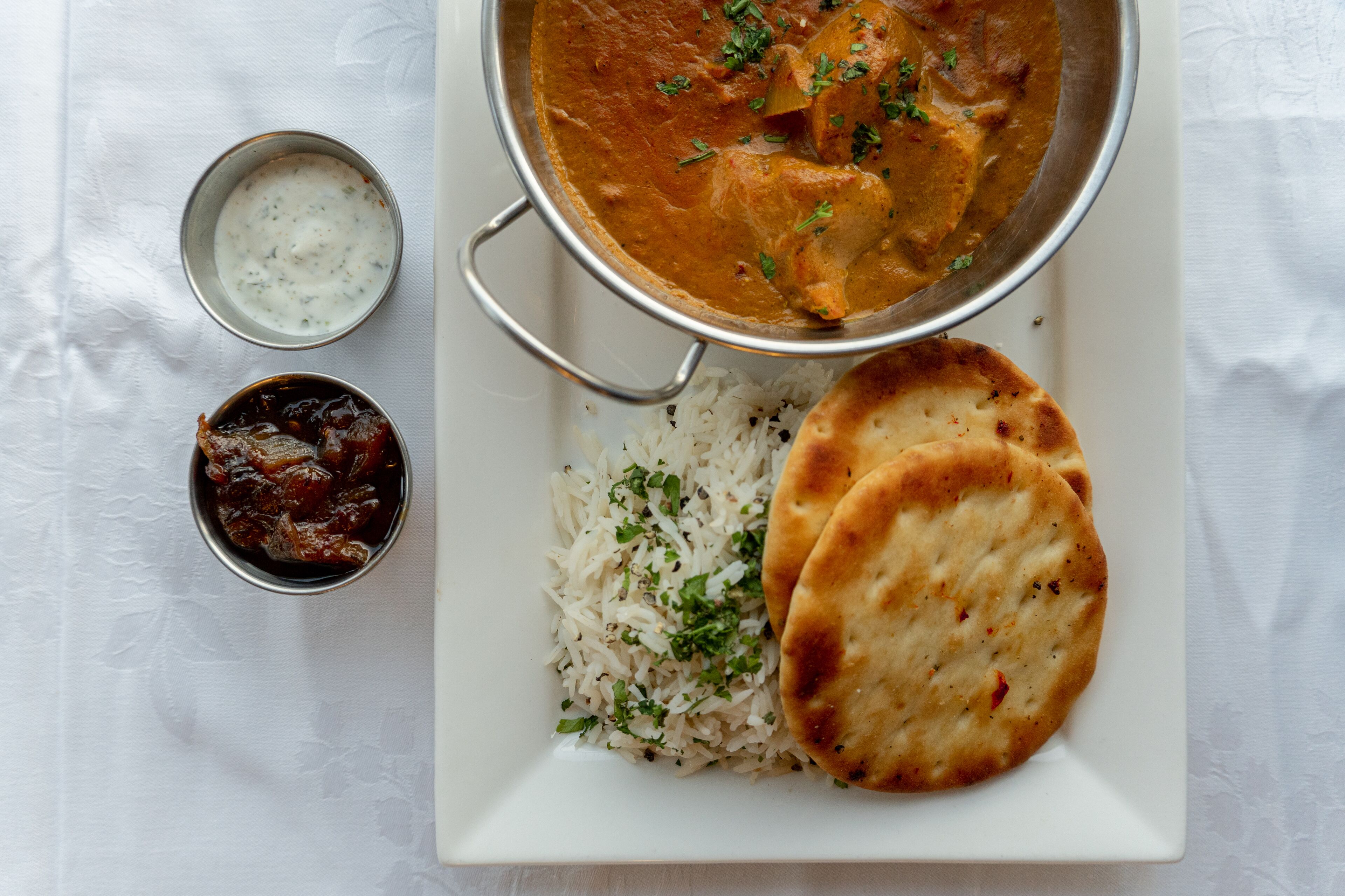 Un plato con arroz, pan naan y un plato de curry, acompañado de dos recipientes pequeños de chutney y raita.