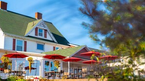 A two-story building with a green roof and red shutters, featuring outdoor seating with red umbrellas. - Auberge Gisele's Inn (Baddeck)