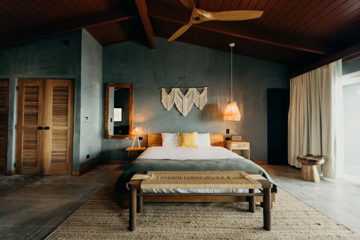 A bedroom with a wooden bed, a ceiling fan, a wall-mounted macrame piece, and a large window with curtains.