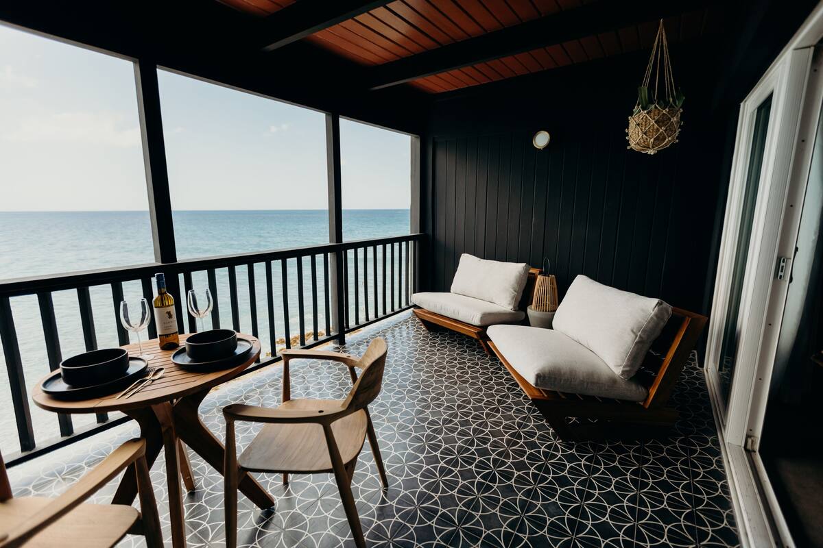 A balcony with a view of the sea, a table with wine glasses and a bottle, and a sofa with cushions.