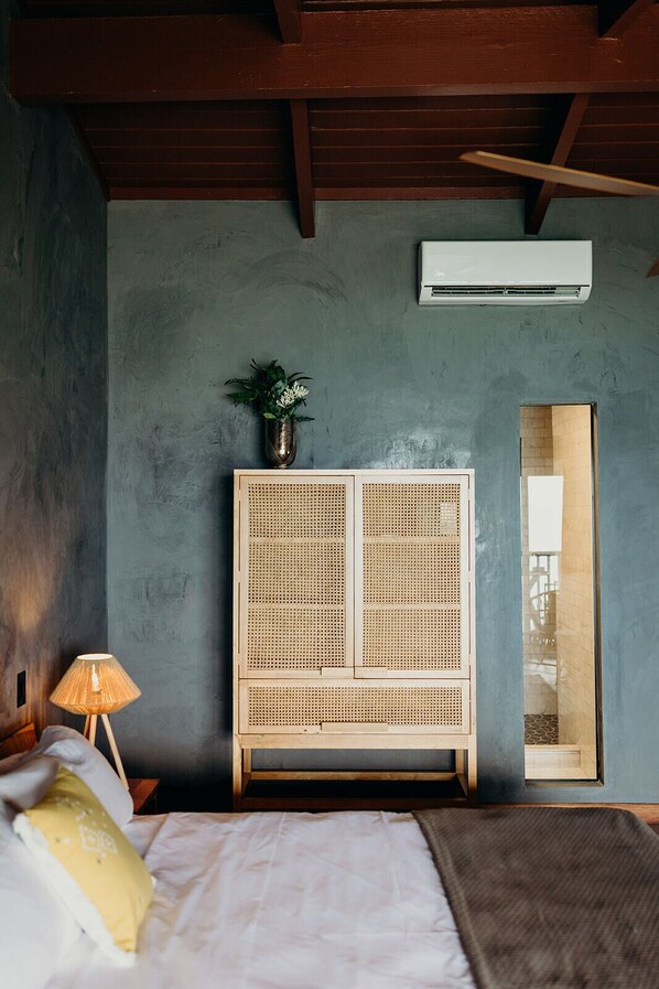 A bedroom with a bed, a bedside lamp, a rattan wardrobe, a potted plant, and an air conditioning unit.