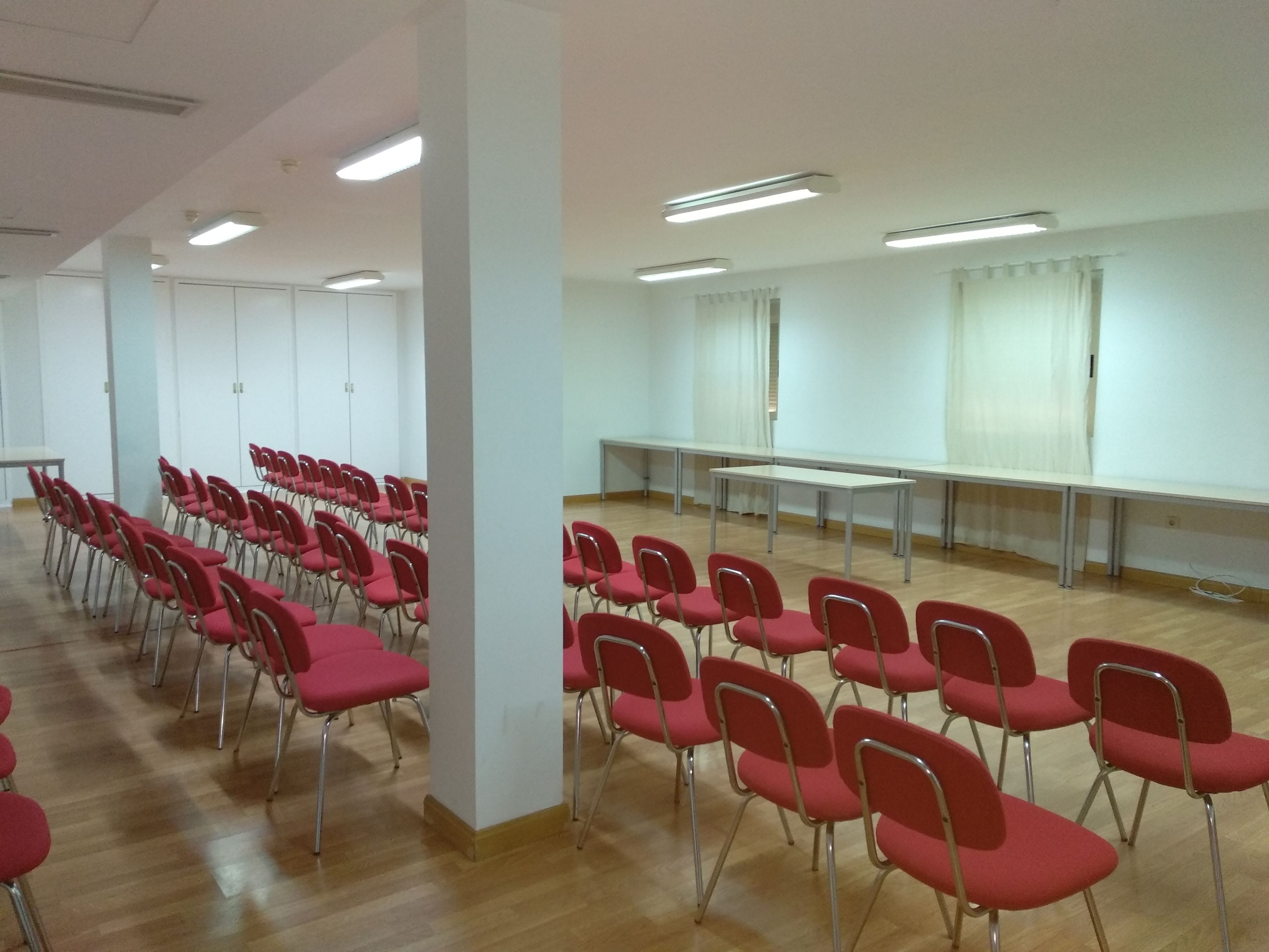 A conference room with rows of red chairs and a long table, white walls, and ceiling lights.