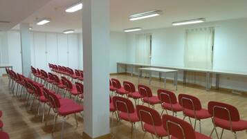 Un salón de conferencias con hileras de sillas rojas y una mesa larga, paredes blancas y luces en el techo.